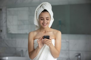 Smiling young woman with white bath towel on head using smartphone 