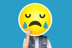 Unrecognizable man hiding his face behind crying sad emoticon emoji over blue background, creative collage with copy space